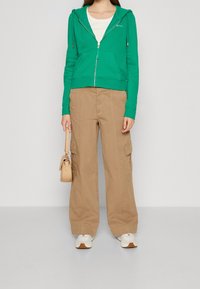 Felpa verde con zip e tasche laterali, abbinata a pantaloni cargo beige larghi. Una borsa beige e sneakers bianche completano l'outfit.