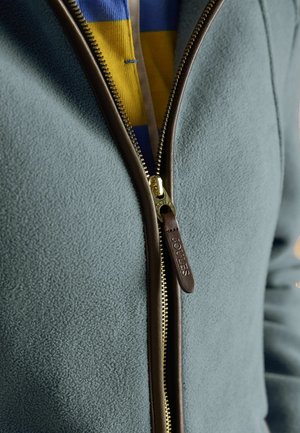 Nahaufnahme einer teilweise zugezogenen petrolfarbenen Fleecejacke mit braunem Lederbesatz und einem darunter sichtbaren gestreiften gelben und blauen Hemd.