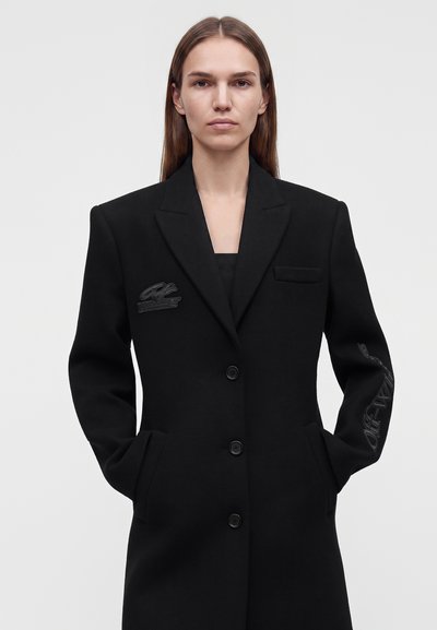 Femme aux longs cheveux bruns porte un manteau noir cintré avec des boutons, des poches latérales et des logos brodés discrets sur la poitrine et la manche droite.