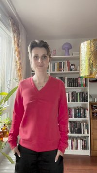 Piros, hosszú ujjú, V-kivágású pulóver, amelynek a mellkasa redőzött, fekete nadrággal viselve, beltérben, könyvespolcokkal a háttérben.