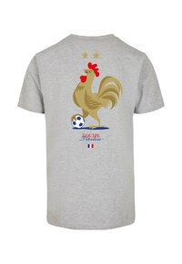 Pilkos spalvos medvilnės marškinėliai su auksinės spalvos gaidžio grafika, turinčiu raudoną žvyną, stovinčiu ant futbolo kamuolio. Viršuje dvi auksinės žvaigždės ir tekstas "Équipe Tricolore."