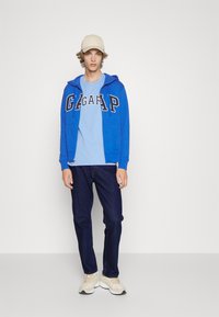Hoodie bleu zippé avec logo, tee-shirt bleu clair en dessous, jean foncé et baskets crème. Porte également une casquette beige.