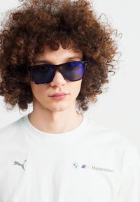 Must have musta prillid siniste klaasidega. Valge T-särk, millel on hall Puma logo ja BMW Motorsport kujundus rinnal. Lokkis juuksed.