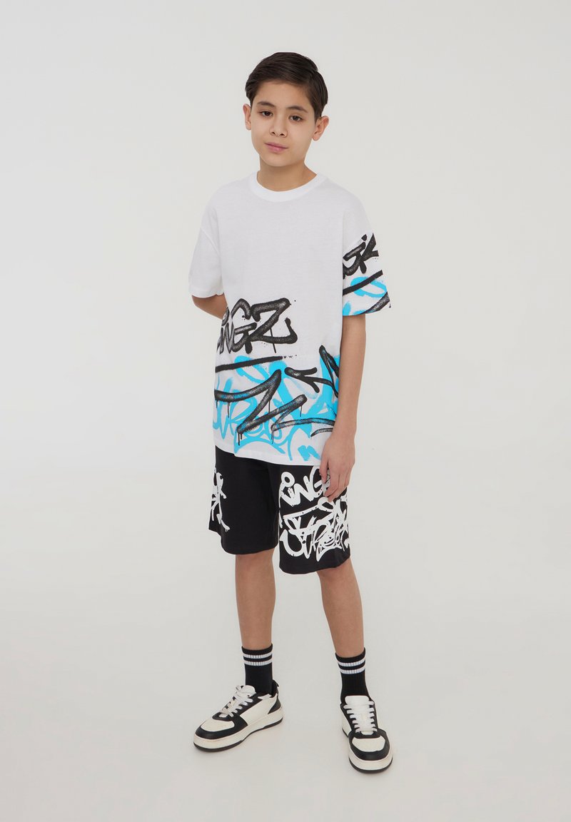 T-shirt bianca a maniche corte con design graffiti nero e blu, abbinata a pantaloni corti neri con motivi graffiti bianchi. Sneakers bianche e nere.