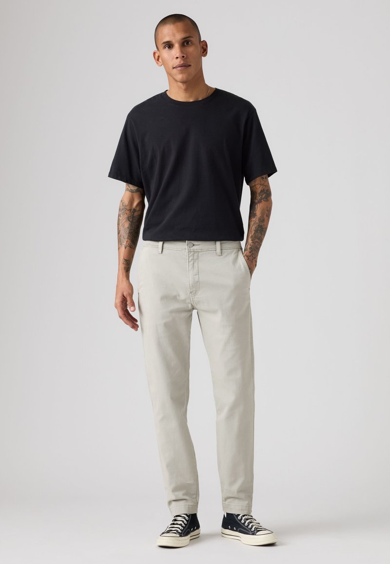 Jeune homme debout, les mains dans les poches, portant un t-shirt noir, un pantalon gris clair, des baskets montantes noires et des tatouages visibles sur les bras.