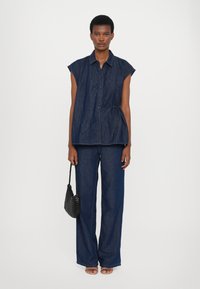 Top de ganga e calças largas em azul escuro, com mangas curtas, botões e um detalhe de cordão, complementados por uma bolsa de mão tecida em preto.