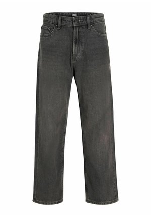 Smilende dreng med krøllet hår iført en enkel hvid t-shirt, løse mørke jeans og hvide sneakers, der står mod en neutral baggrund.