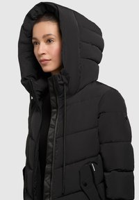 Chaqueta de plumas negra con una capucha grande acolchada, cierre con cremallera en la parte delantera y dos bolsillos laterales, hecha de una tela suave y ligera.