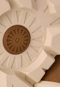 Suola in gomma beige con superficie testurizzata, caratterizzata da un motivo a raggi e logo circolare marrone centrale impresso con la scritta “UGG”.