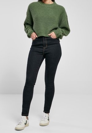 Pull en maille verte à texture côtelée, associé à un jean taille haute foncé. Le modèle porte des baskets blanches avec des accents verts.