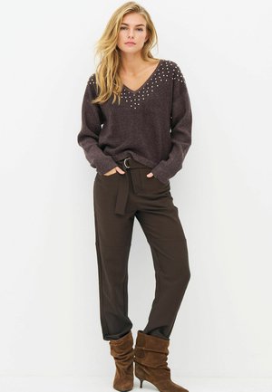 Woman wearing a dark brown sweater with pearl details, dark brown belted pants, and slouchy brown suede boots, standing against a white background.