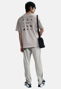 Camiseta gris de manga corta con logotipos impresos en la parte trasera, combinada con pantalones gris claro con detalles negros y un pequeño bolso cruzado negro.