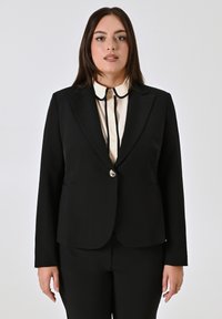 Schwarzer Blazer mit Reverskragen, Einzelknopfverschluss und zwei vorderen Taschen. Getragen über einem cremefarbenen Hemd mit kontrastierendem Kragen.