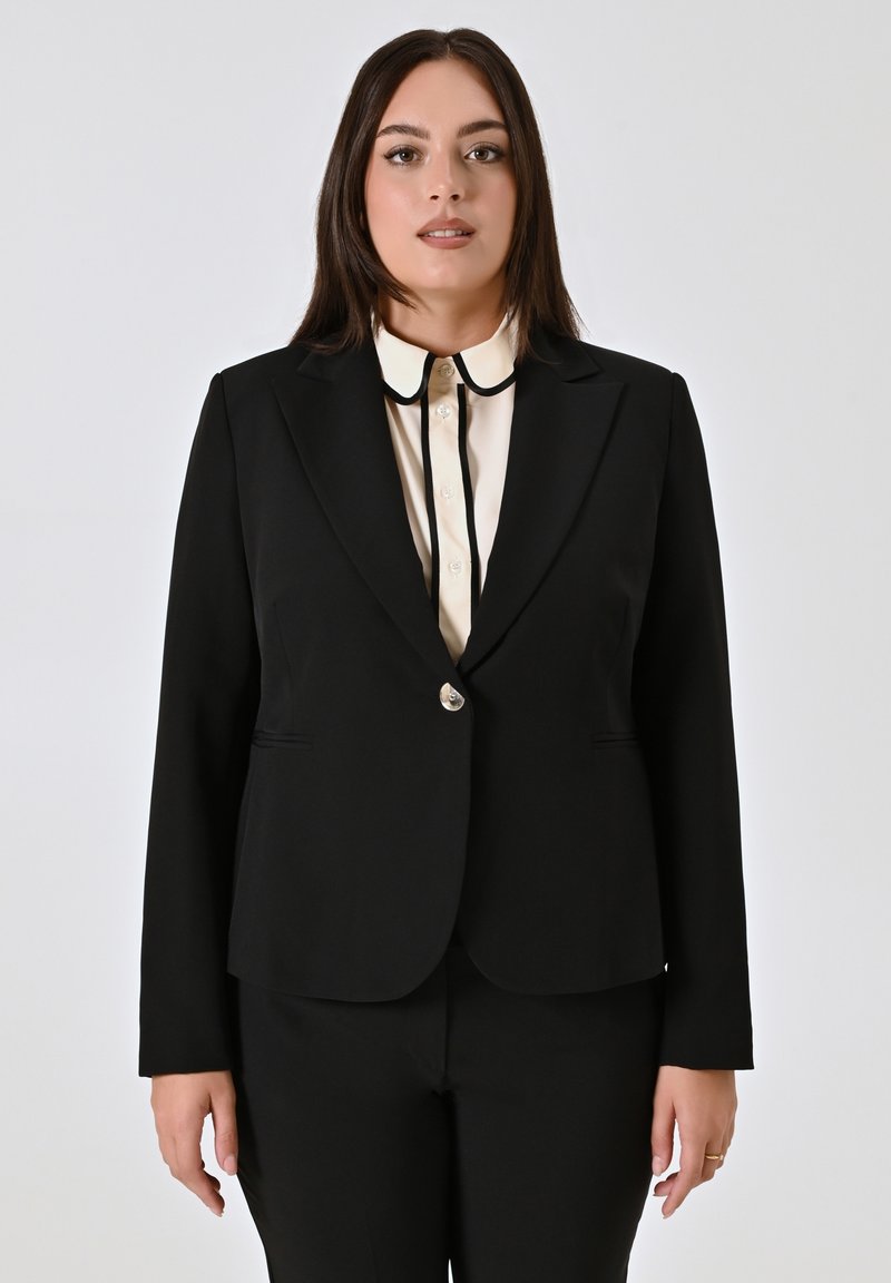 Schwarzer Blazer mit Reverskragen, Einzelknopfverschluss und zwei vorderen Taschen. Getragen über einem cremefarbenen Hemd mit kontrastierendem Kragen.