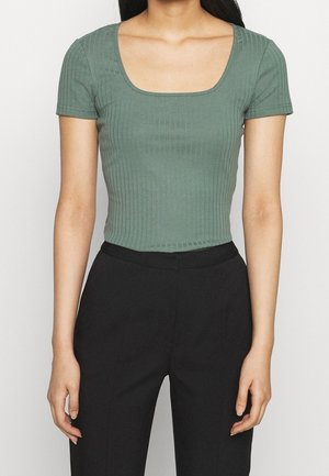 Vrouw draagt een aansluitende groene geribbelde top met korte mouwen en een vierkante halslijn en hoge zwarte broek, tegen een effen achtergrond.