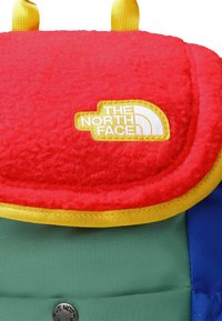 North Face mugursoma ar sarkanu, pūkainu augšējo vāku, dzelteniem akcentiem un zaļu pamatni ar zilu sānu paneli. Logotips ir izteikti redzams.