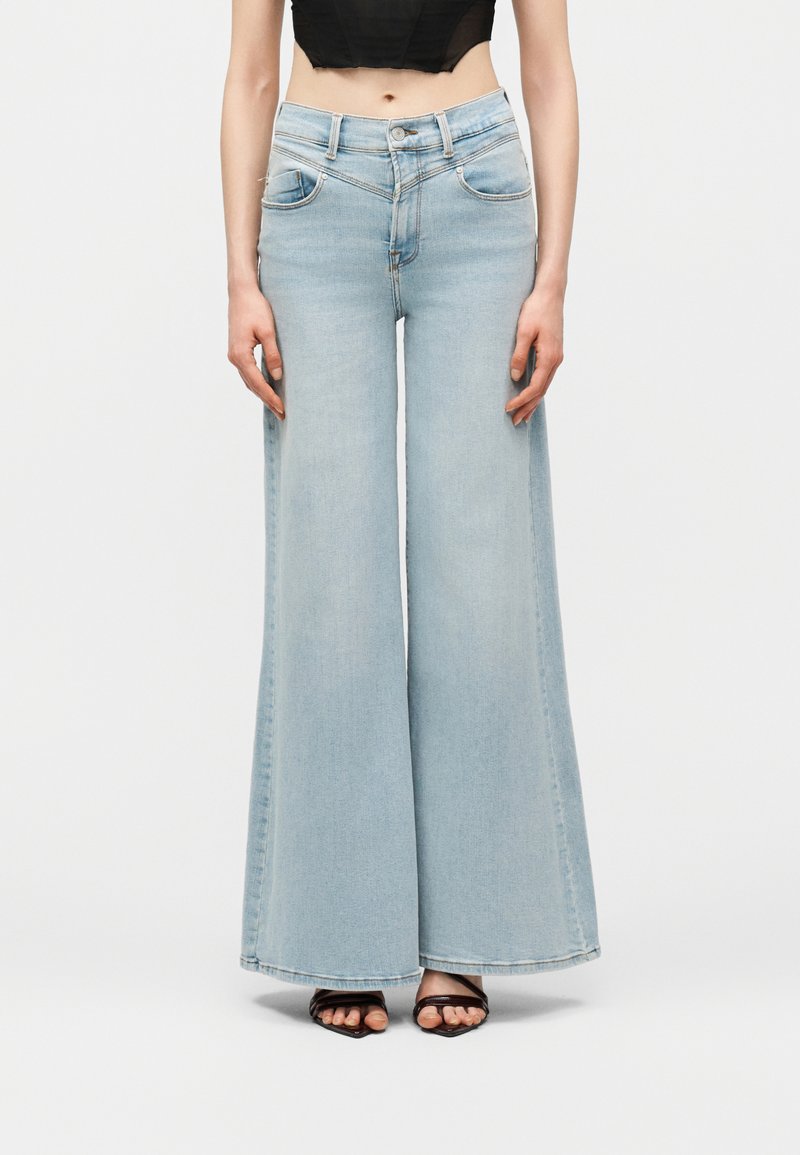 Γυναίκα με ανοιχτόγαλανά τζιν ψηλόμεσα παντελόνια με φαρδιά μπατζάκια, μαύρο κοντό τοπ και μαύρα σανδάλια ανοιχτής μύτης, μπροστά σε λευκό φόντο.