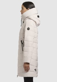 Veste blanche rembourrée avec un col haut et une capuche, présentant des coutures verticales, des poches latérales et un patch de logo sur la manche.