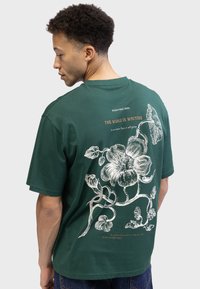 Dunkelgrünes Baumwoll-T-Shirt mit einem weißen Blumenmuster auf der Rückseite, ergänzt durch Text und Akzente in einer helleren Farbe. Kurze Ärmel und Rundhalsausschnitt.