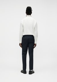 Personne aux cheveux sombre tressés, vue de dos, portant une chemise blanche à manches longues, un pantalon foncé sur mesure et des chaussures habillées noires.