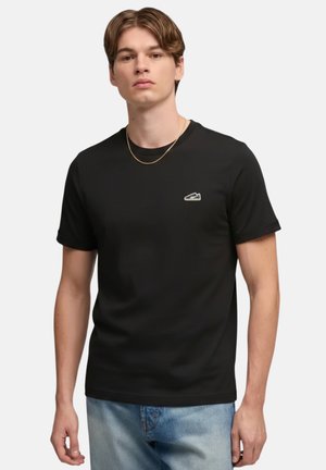 Giovane uomo che indossa una t-shirt nera a maniche corte con un piccolo logo sul petto e jeans azzurri, in piedi davanti a uno sfondo neutro.