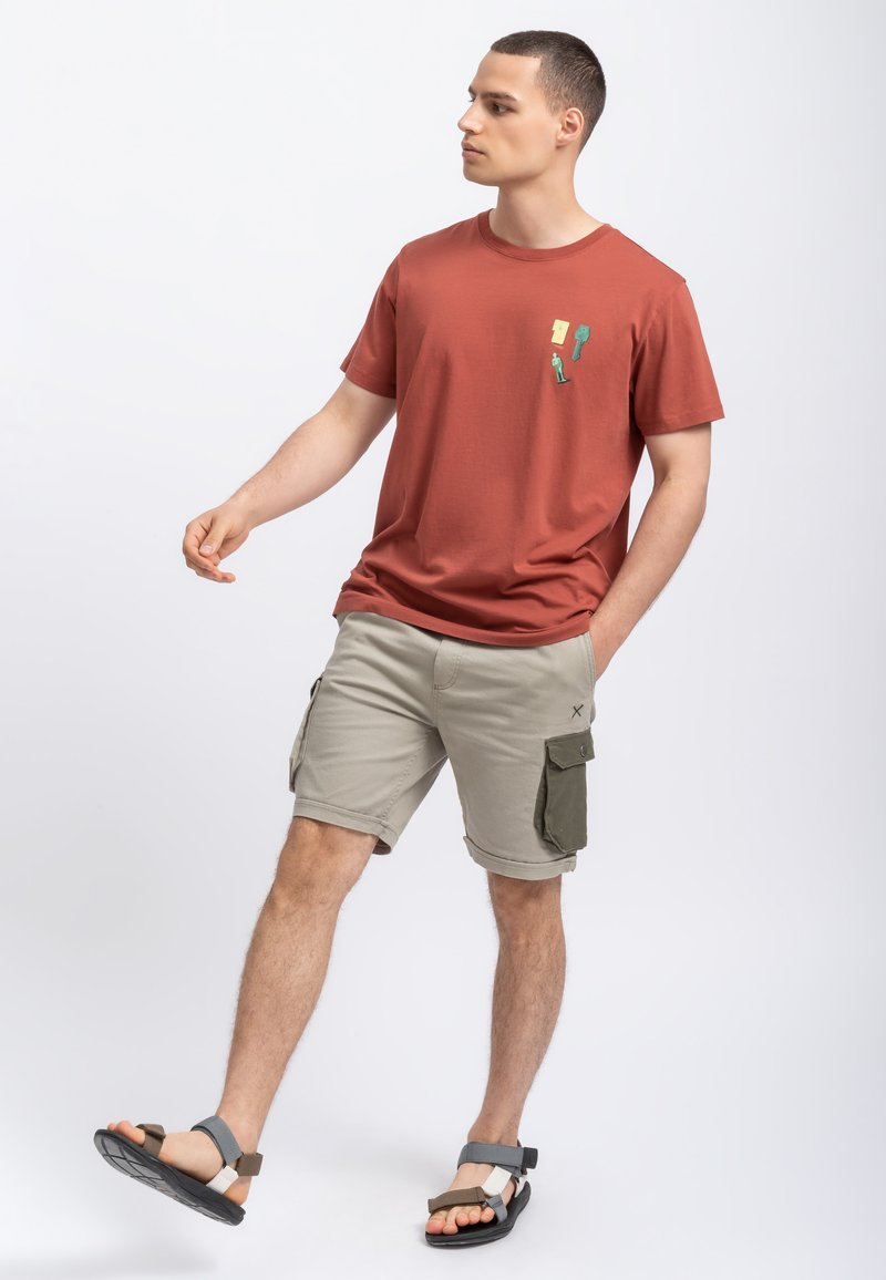 T-shirt in cotone color ruggine con grafica tasca, abbinata a pantaloni cargo beige con accenti grigi e verdi, e sandali neri.