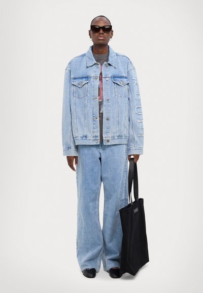 Chaqueta de mezclilla azul claro con dos bolsillos en el pecho, combinada con jeans de mezclilla a juego. Un bolso tote negro y unas gafas de sol oscuras completan el conjunto.
