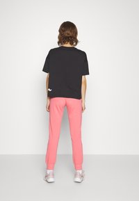 Schwarzes T-Shirt mit kurzen Ärmeln und einem weißen Logo auf dem Rücken, kombiniert mit rosa Jogginghosen, abgerundet mit hellen Sneakern.