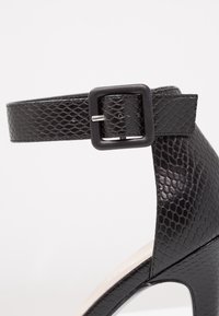 Sandale noire à talons hauts réalisée dans un matériau texturé, ressemblant à de la peau de reptile. Elle est dotée d'une large bride de cheville avec une boucle carrée noire et présente une forme lisse et arrondie.