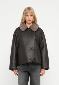 Sort læderjakke med en brun shearling-krave, der har en knaplukning foran og brede ærmer, parret med grå jeans.