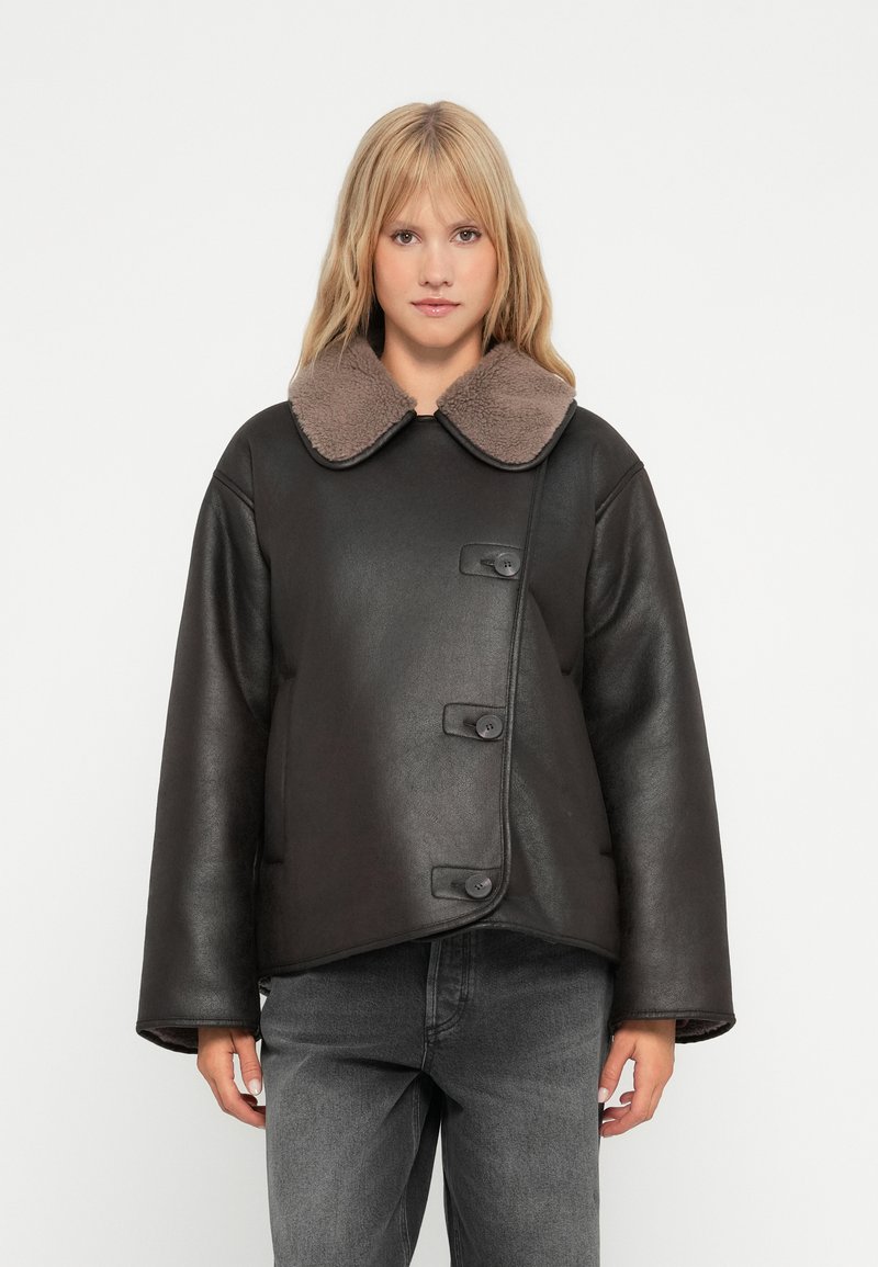 Sort læderjakke med en brun shearling-krave, der har en knaplukning foran og brede ærmer, parret med grå jeans.