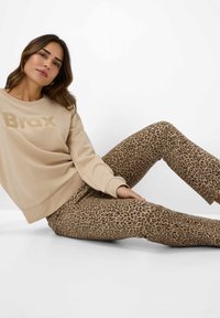 Beige sweatshirt met het "Brax" logo. Aansluitende broek met een bruin panterprint. Model zittend, waarbij beide kledingstukken tegen een witte achtergrond worden getoond.