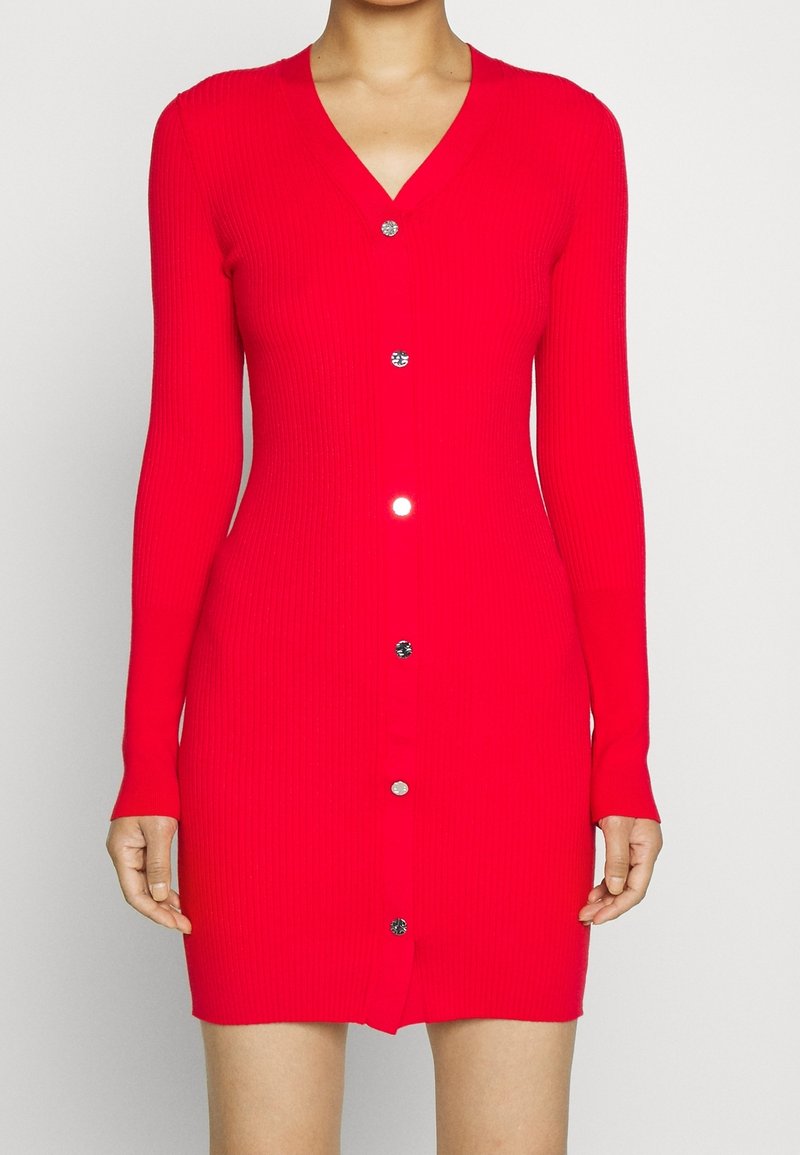 Femme portant une robe cardigan rouge cintrée à manches longues avec des boutons ronds argentés, debout devant un fond clair uni.