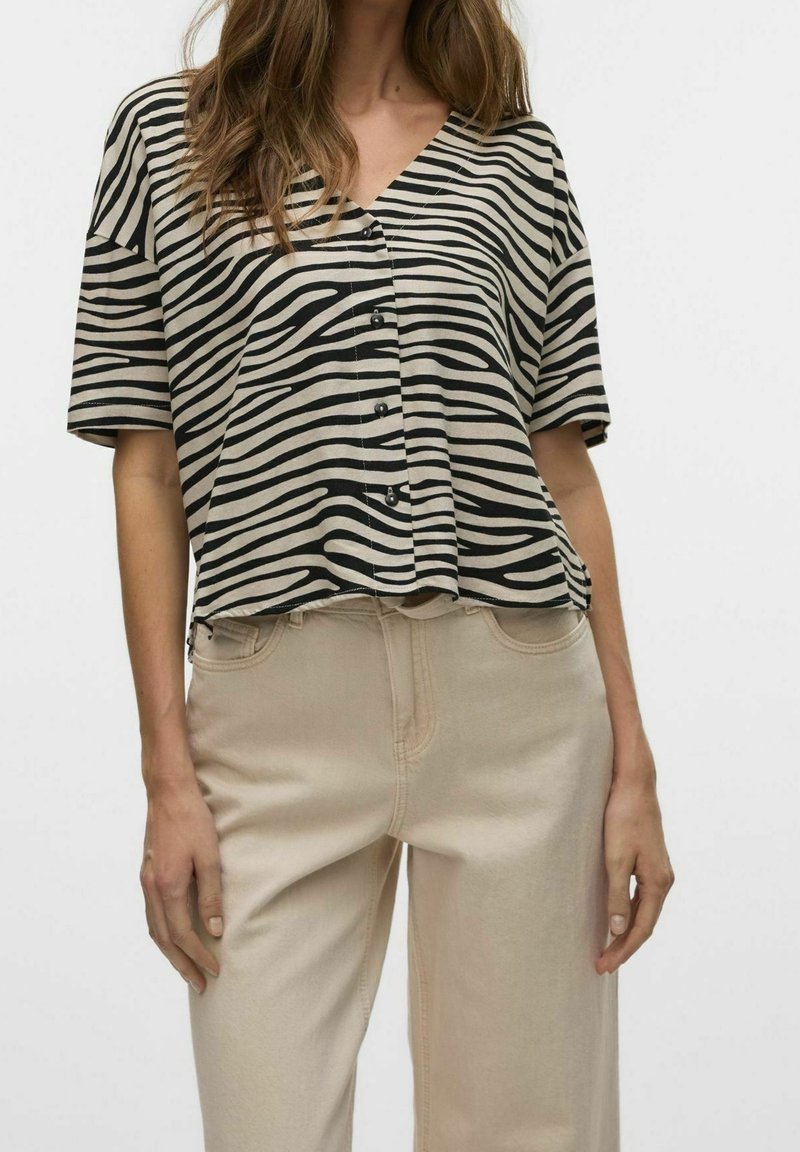 Haut à manches courtes et décolleté en V beige avec des rayures zèbres noires, doté de boutons sur le devant et d'une coupe décontractée. Associé à un jean beige clair.