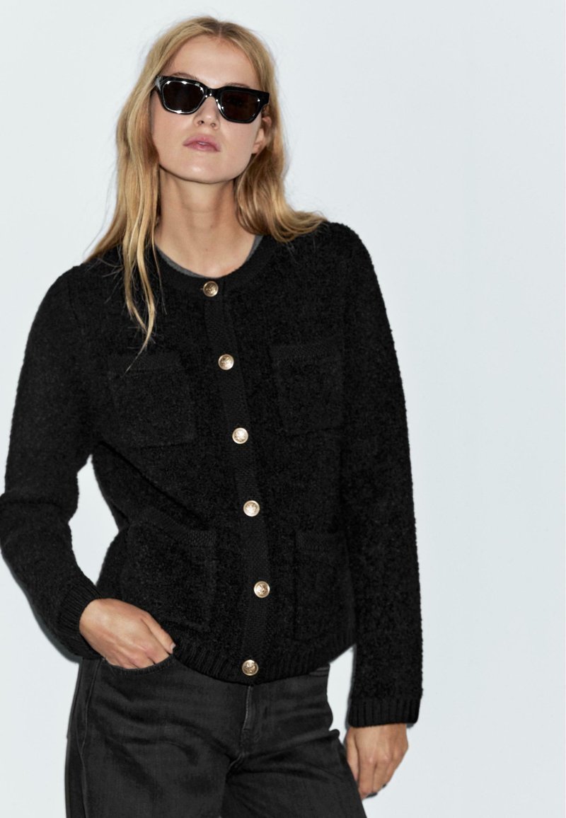 Donna bionda che indossa un cardigan nera con bottoni e occhiali da sole, con una mano in tasca, su uno sfondo chiaro e uniforme.