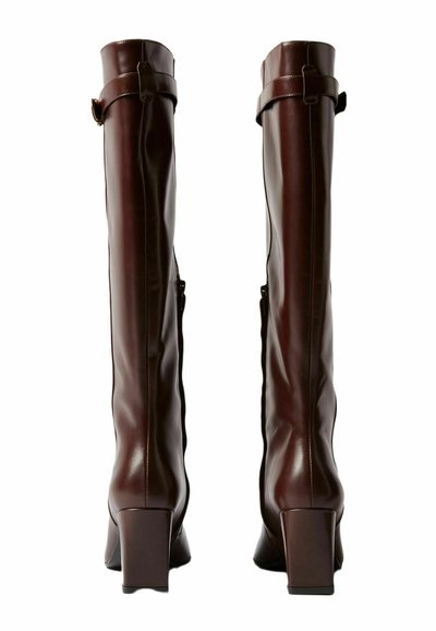 Botas de cuero marrón hasta la rodilla con un acabado brillante, tacones cuadrados y cremalleras laterales. Presentan una tira de cinturón en la parte superior.