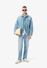 Chaqueta de denim azul claro superpuesta a un chaleco de denim a juego, combinada con jeans azul claro y zapatillas de crema. Sosteniendo un bolso amarillo plegado.