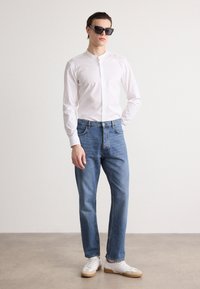 White long-sleeve button-up shirt, blue jeans, and white sneakers with tan soles. Model wearing oversized black sunglasses, standing against a light background.