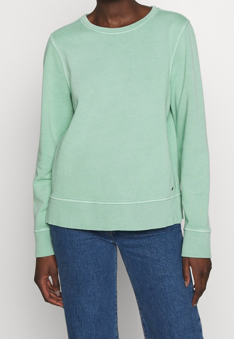 Personne portant un sweat-shirt vert clair à manches longues et un jean bleu, debout devant un fond blanc uni.