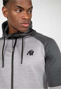 Siva i crna zip-up dukserica s suptilnim heather uzorkom, s logom gorile na prsima i podesivom kapuljačom.