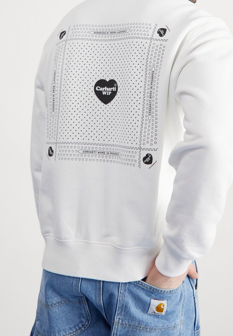 Sweat-shirt blanc avec logo Carhartt WIP en forme de cœur noir et petits motifs de cœurs au dos, porté avec un jean en denim bleu.
