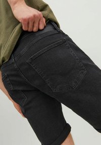 Persona con pantalones cortos de mezclilla negros arremangados y una camiseta verde oliva, levantando ligeramente la camiseta por detrás.