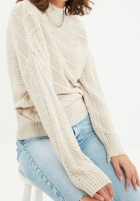 Pull en tricot crème avec ourlet côtelé et manches larges ; présente un motif texturé et un col rond. Associé à un jean bleu clair.