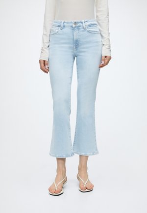 Personne portant un jean flare court bleu clair, un haut blanc à manches longues et des sandales blanches, debout devant un fond uni clair.