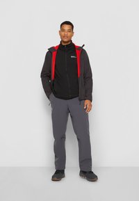 Zwarte zip-up jas met een rode capuchon, gecombineerd met een zwarte basislaag en grijze broek. De schoenen zijn zwart met bruine accenten.