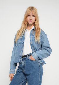 Ung kvinne med langt blondt hår iført lys blå denimjakke og jeans over hvit skjorte, står med den ene hånden i lommen mot en enkel bakgrunn.