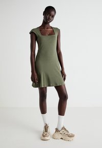 Vestido verde-oliva de manga curta com decote quadrado, feito de tecido suave, design fluido, combinado com meias claras e ténis robustos.