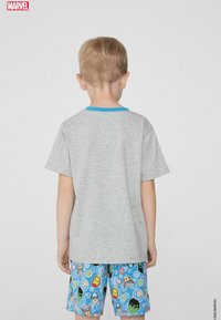 Graues Kurzarm-T-Shirt mit blauem Kragen, kombiniert mit bunten Shorts mit Superhelden-Mustern auf hellblauem Hintergrund.