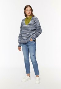 Gebreide blauw- en grijsgestreepte trui over een groene overhemd met kraag, gecombineerd met lichtblauwe skinny jeans en witte sneakers.