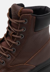 Bota de senderismo de cuero marrón con superficie texturizada, cuello acolchado, ojales de metal y cordones marrones. Presenta una suela de goma negra y costuras en contraste.